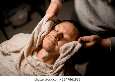 Barber covering client's face with hot towel before giving him shave at barbershop. Procedure to make skin and hair smoother for easier shaving - Powered by Shutterstock