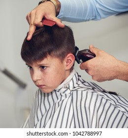 Barber is doing haircut to Caucasian boy using hair clipper in barbershop. 
