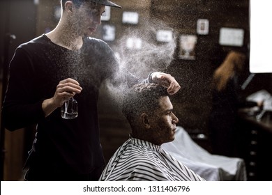 The Barber Dressed In A Black Clothes Is Doing The Hair Styling To The Stylish Black-haired  Man Sitting In The Armchair In A Barbershop