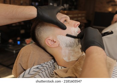 Barber Man is shaving client with a vintage straight razor in a barbershop. Shaving cream, foam. Classic shave by stainless steel straight edge razor