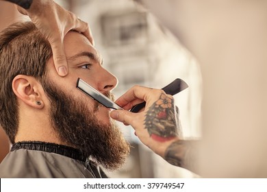 Barber with old-fashioned black razor shaving bearded man 