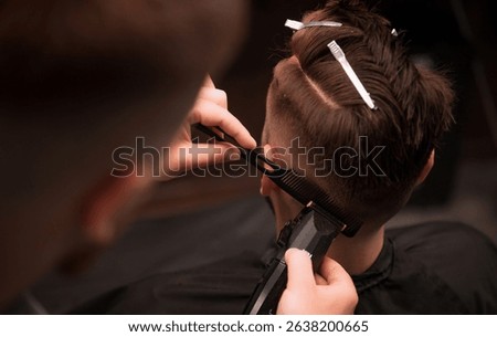 Barber precisely trims the side of a man's head with electric clippers and a comb. Hair is sectioned with clips, showcasing detailed grooming in a professional barbershop setting.