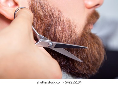 Barber with scissors shaving bearded man