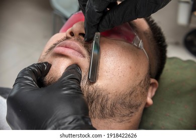 A barber shapes an asian man's beard and sideburns with a stainless razor. Professionally done facial hair styling at a barbershop or salon.
