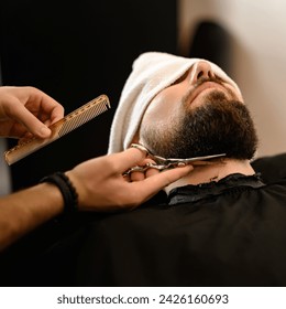 A barber stylist trims the beard of a Caucasian man, whose face is covered with a towel, with scissors - Powered by Shutterstock