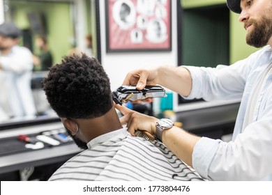 Barber Trim Hair With Clipper On Young Unshaven Black Man In Barbershop Studio. Professional Hairdresser Cut Hair With Electric Shearer Machine On African Guy.