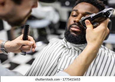 Barber Trim Hair With Clipper On Young Unshaven Black Man In Barbershop Studio.Professional Hairdresser Cut Hair With Electric Shearer Machine On African Guy.