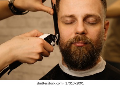 Barber trimming beard of male customer with electric razor at barbershop. Man with beard and moustache getting stylish trendy hair shaving, with hair clipper and comb. Close up. - Powered by Shutterstock