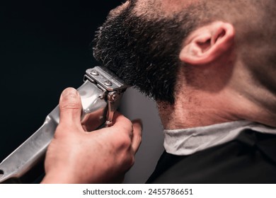 A barber trims a man's beard. The barber is holding a silver-colored electric beard trimmer. The man is sitting in a chair and is relaxed. - Powered by Shutterstock