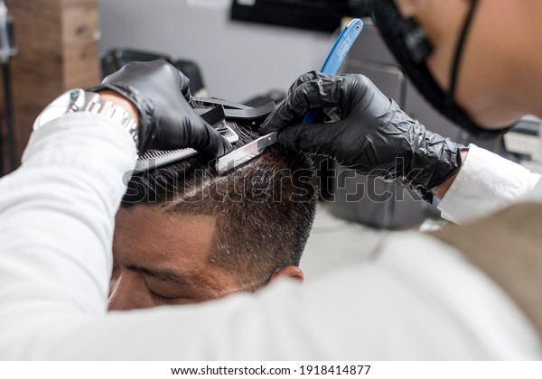 A barber wearing gloves uses a shaving knife\
to create a hard part on an asian man\'s scalp. Classic and trendy\
haircut style. At a\
barbershop.