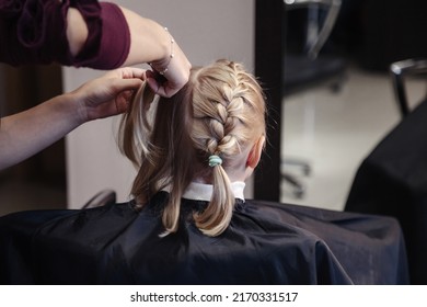 Barber woman braids pigtails, makes fashionable pretty hairstyle for cute little blond girl child in barbershop. Hairdresser makes hairdo for young baby in barber shop. Concept hairstyle and beauty