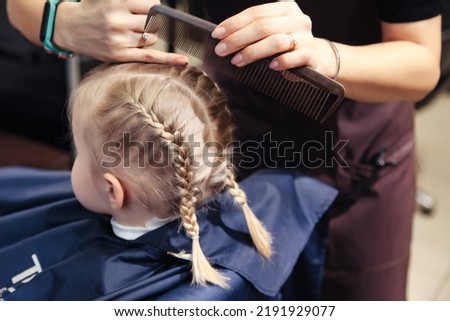Barber woman make fashionable hairstyle for cute little blond girl child in modern barbershop. Hair salon, Hairdresser makes hairdo braids pigtail for young baby in barber shop. Copy space