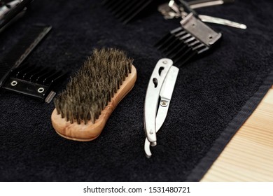 Barber Working Tools In A Barbershop. Natural Base Brush, Comb, Hazardous Razor, Shaving Machine With Nozzles Are All On A Black Terry Towel For Client's Haircut. Hair And Beard Care And Beauty