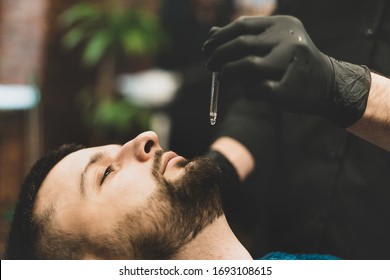 Barbershop. The client in the master’s chair in the barbershop, the barber applies oil and cosmetics to the client’s beard. Male beauty shop. Healthy lifestyle and beauty