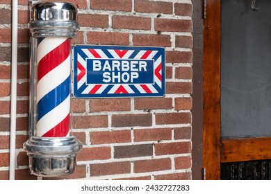 A barbershop pole with red, blue, and white stripes moving around on the outside of a men's shop. The vintage pole is attached to a brown brick wall. There's a sign with Barber Shop in white letters. - Powered by Shutterstock