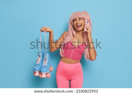 Barbie style girl in pink clothes and pink wig talking on phone and smiling, holding blue roller-skates, posing on blue background, personal style concept, copy space