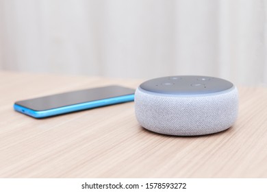 Barcelona, Spain - 03 of december of 2019: amazon echo dot third generation, white, with a blue smartphone out of focus, on a light wooden table