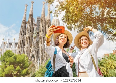 BARCELONA, SPAIN - 11 JULY 2018: Young Girls Friends Making Selfie Photo On Her Smartphone In Front Of The Famous Sagrada Familia Catholic Cathedral. Travel In Barcelona Concept