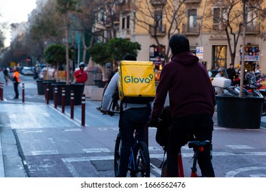 Barcelona, Spain - 20 February 2020: Young Riders Working For Glovo Delivery Service Riding Bike In The City In The Evening. Food Delivery Plays A Mayor Role In The New Gig Economy