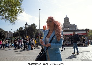 Barcelona, Spain - April 12, 2022: Older Woman With Large Eyes Made Up And Hair With Pink Tones, Dressed In A Sky Blue Jacket. She Stands In The Middle Of A Busy Pedestrian Sidewalk 