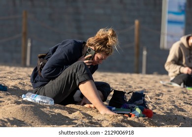 Barcelona, Spain - Jan 08, 2022: A Young Woman, Barefoot, Sitting On The Sand Of Barceloneta Beach, In The Afternoon, Talking To Someone On Her Cell Phone