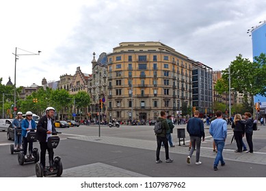 Barcelona, Spain - May 5, 2018:  People Ride Electric Segway Scooters Through Intersection Of Passeig De Gràcia A High End Shopping Area.  Casa Battlo Is To The Left Of The Building On The Corner.