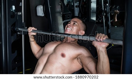 A bare chested man in the middle of doing intense incline barbell bench presses on a Smith Machine at a gym or fitness center. In the zone and motivated doing chest muscle training.