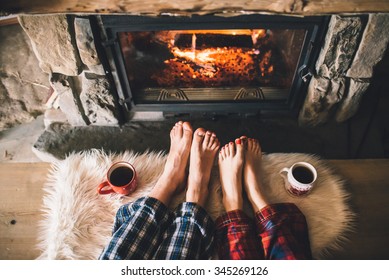 Bare couple feet by the cozy fireplace. Man and Woman relaxes by warm fire with a cup of hot drink and warming up her feet. Close up on feet. Winter and Christmas holidays concept.