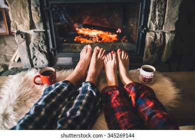 Bare couple feet by the cozy fireplace. Man and Woman relaxes by warm fire with a cup of hot drink and warming up her feet. Close up on feet. Winter and Christmas holidays concept.