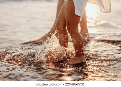 The bare feet of an adult woman and a girl, a child in white dresses are splashing in the ocean at sunset. Water drops sparkle in the sun - Powered by Shutterstock