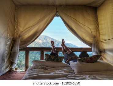 Bare feet of a couple lay down on the bed by mountains view in the morning. View from inside a tent. Dark foreground photo. Love is all around.
