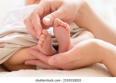 Bare Feet Of A Newborn Baby On Mother's Palm. Women's Hands Gently Hold And Touch Little Toes. White Blanket On The Background. Lifestyle. Concept Of Motherhood, Childhood, Child Care. Selective Focus