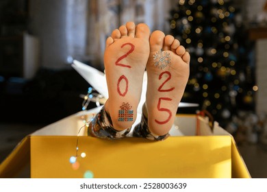 bare feet with the numbers 2025 written on them peek out from a large cardboard box against the backdrop of a Christmas tree. festive atmosphere on New Year's Eve. Cheerful childhood