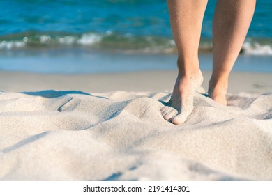 Bare female feet on a sandy beach with blue sea in the back on a beautiful sunny summer day. Concept for holiday, vacation, relaxation, mental health, joy, recreation.