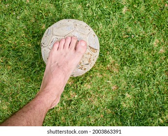 Bare Foot Of A Man Standing On An Old Foot Ball, On The Grass, No Faces Shown