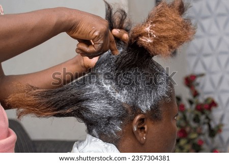 Bare hands of an African Nigerian stylist, applying relaxer cream to the natural long hair of a woman or female customer at a beauty salon in Nigeria
