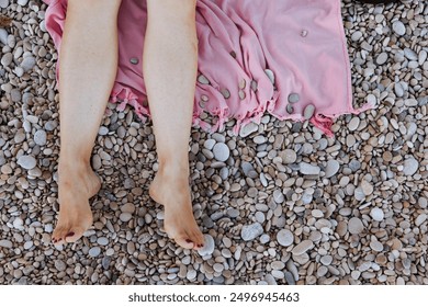 Bare legs resting on a pink blanket placed on a pebbled beach, creating a relaxed and tranquil scene. - Powered by Shutterstock