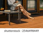 Bare legs of unrecognizable woman in a robe on a deck chair basking in the sun. The wooden decking is adorned with fruits and books, creating a relaxing outdoor atmosphere.