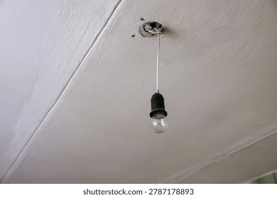 A bare light bulb hanging from a ceiling fixture. The ceiling is plain and white, showing signs of wear. The light bulb is unadorned and exposed.