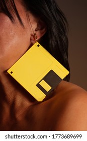 Bare Shouldered Female (face Obscured) With Black Hair Wears Bright Yellow 3.5 Inch Floppy Diskette As An Earring In 1980s Or 1990s Retro Fashion