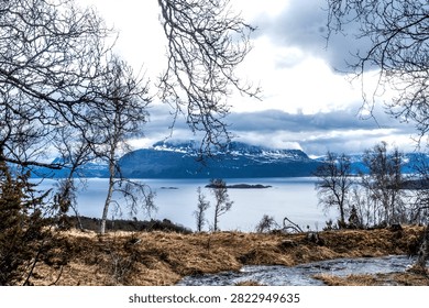 Bare trees and a small flowing stream frame a calm fjord and a snow-covered mountain beneath dramatic clouds in Northern Norway.