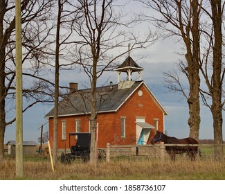 Bare Winter Trees With Horse And Buggy In The Foreground And Old Red Brick School In The Background Of This Rural Scene. 