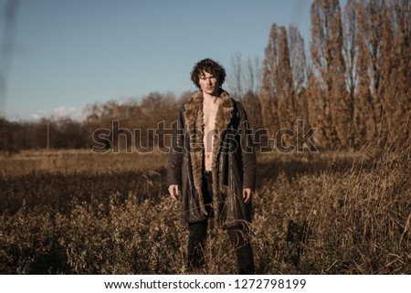 a bare-chested boy wears a fur coat standing in a wild meadow. He looks at you