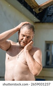 A bare-chested man cools off in the heat under a stream of summer rain flowing down from a roof