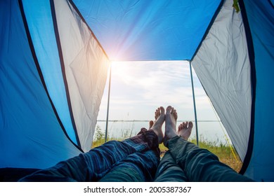 Bared legs of man and woman stretched out of tourist tent. View on river bank with green grass and blue sky on sunny summer day. Crossed barefoot of lovers touching. Family tourism, honeymoon.