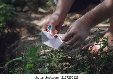 A barefoot adult man lights a paper boat. Hands holding a white children's origami boat and a burning match. Summer time. Selective Trick. Outside the room.