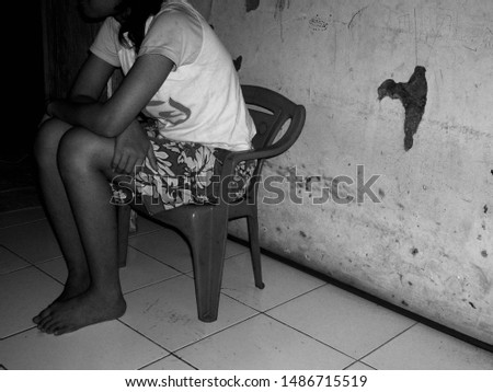 A barefoot Asian teenage girl sits in an undersized plastic chair in a dilapidated dark grimy room with peeling plaster walls.