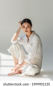 Barefoot asian woman in white clothes sitting on grey background