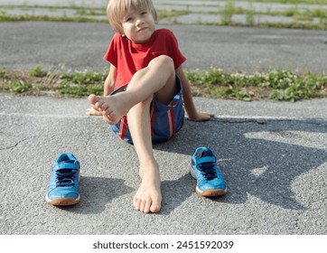 barefoot boy sits on the asphalt and children's blue sneakers stand nearby. Take off your shoes in summer and cool your feet after sports - Powered by Shutterstock