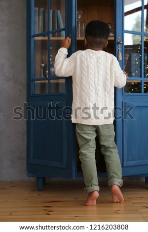 A barefoot boy in a white knitted sweater stands on on his toes near the  room decorated for New Year and Christmas